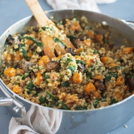 A pot filled with risotto, containing rice, mushrooms, spinach, and pieces of butternut squash. A wooden spoon is partially submerged in the dish.