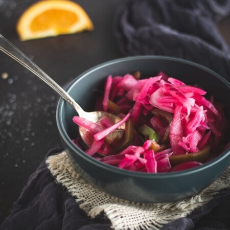 A bowl of bright pink pickled vegetables with a spoon, placed on a woven mat and dark fabric. A slice of orange is in the background.