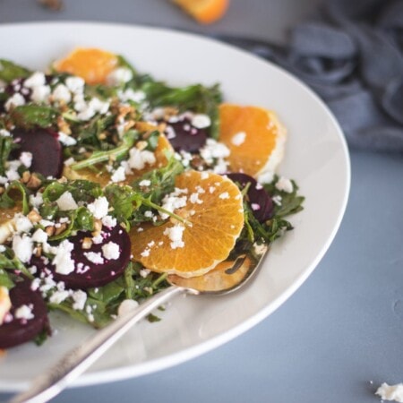 A salad with orange slices, beetroot, greens, nuts, and crumbled cheese on a white plate. A fork is resting on the plate. Orange slice and gray cloth are in the background.