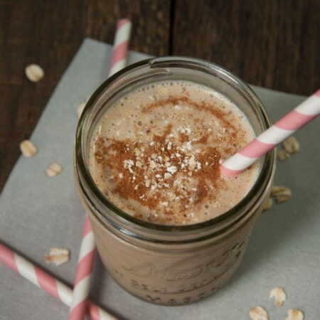 A jar of smoothie topped with cinnamon and oats on a gray surface. Pink-striped straws are placed in and around the jar. Scattered oats are visible on the surface.
