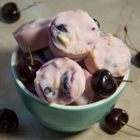 A bowl of pink frozen yogurt bites with cherries and nuts, surrounded by fresh cherries on a beige surface.