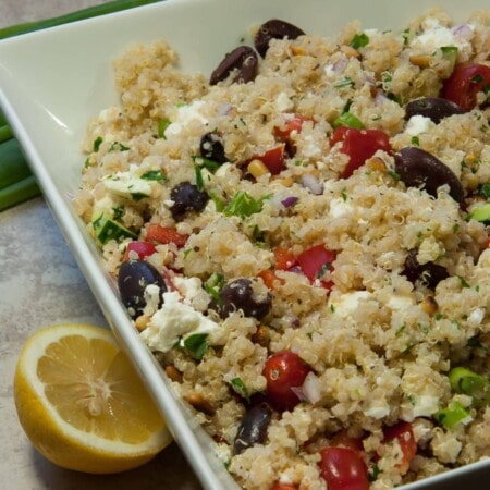 A dish of quinoa salad with cherry tomatoes, black olives, feta cheese, and herbs, garnished with a lemon half in a white square bowl.