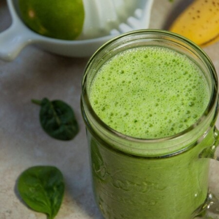 A glass jar filled with green smoothie on a table with a lime juicer, a banana, and spinach leaves nearby.
