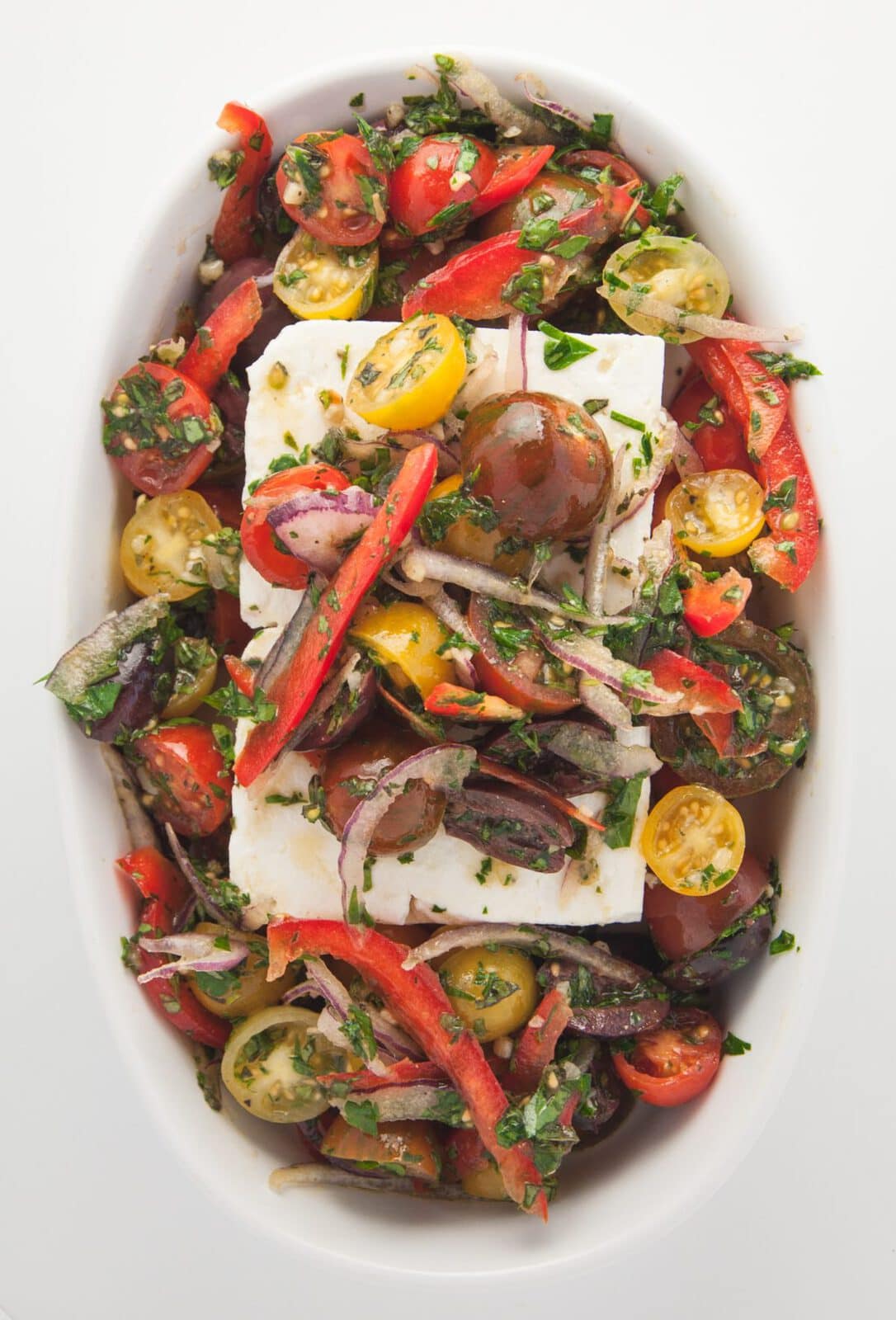A colorful dish with unbaked cherry tomatoes, red onion, sliced peppers, and herbs surrounding two blocks of white cheese on a white oval plate.