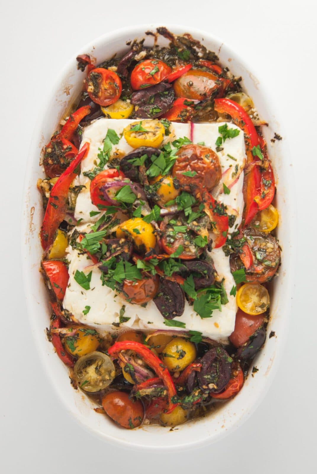 Baked feta cheese surrounded by roasted cherry tomatoes, olives, and sliced red peppers, garnished with chopped parsley in a white oval dish.