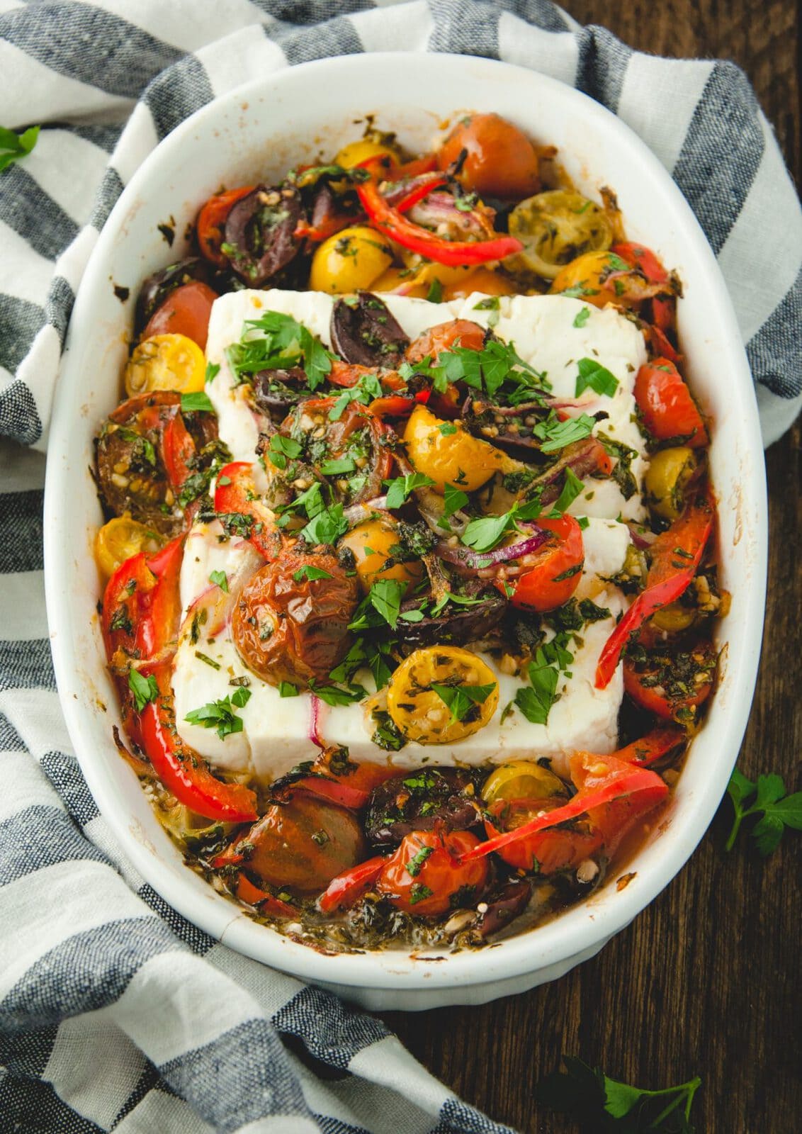 White dish with baked feta topped with tomatoes, herbs, olives and peppers