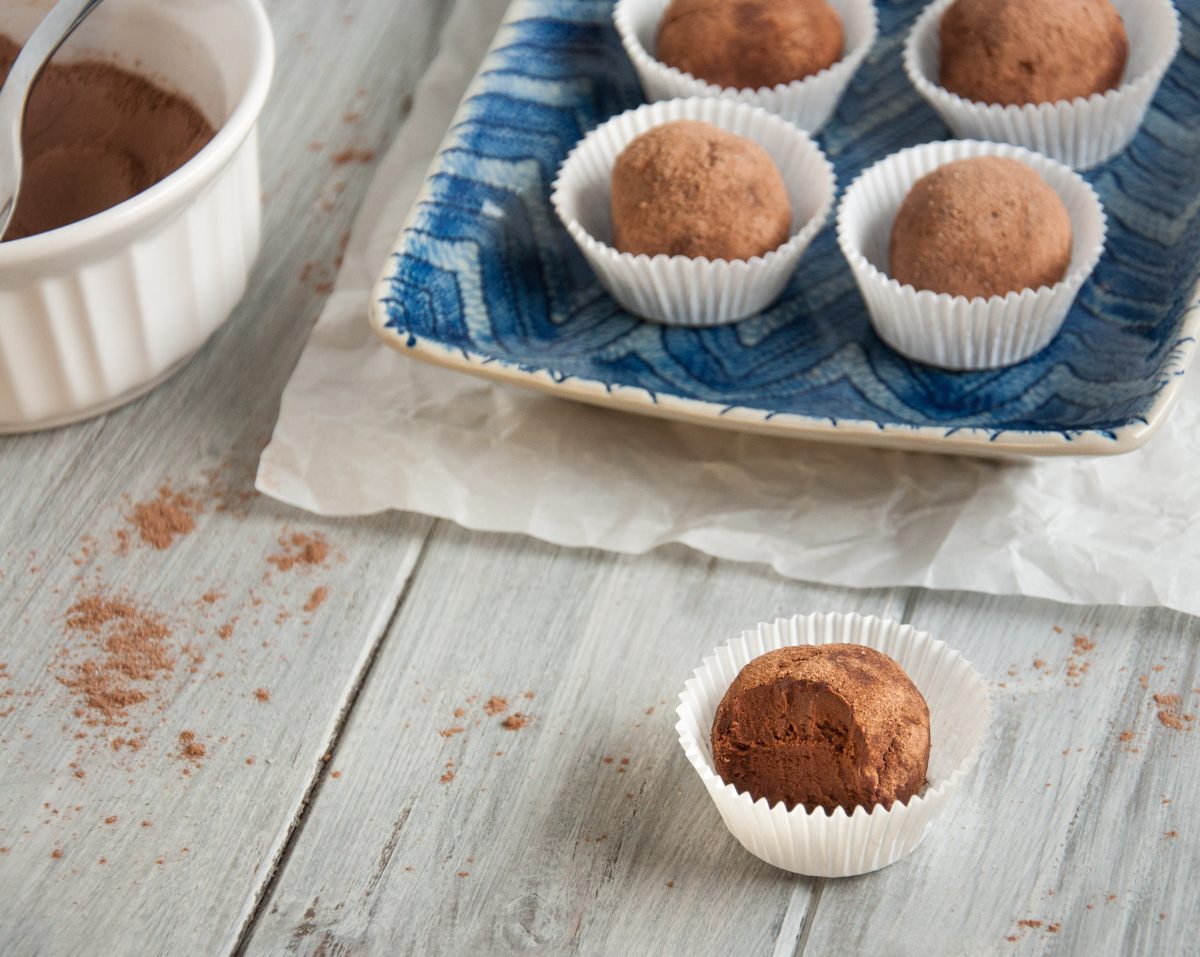Chocolate truffles in white paper cups rest elegantly on a blue plate set upon a wooden table, accompanied by a bowl of cocoa powder.