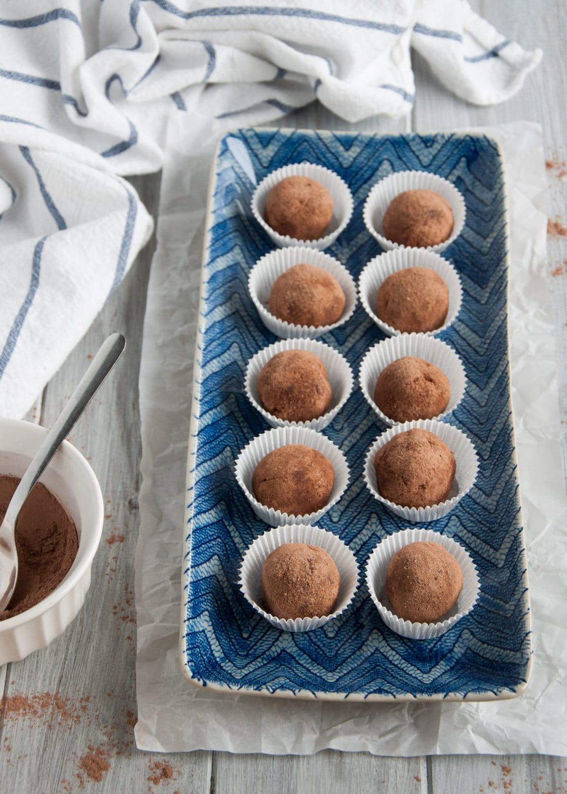 A rectangular blue plate holds ten delectable avocado chocolate truffles, each nestled in a white paper cup. Nearby, a bowl of cocoa powder and a spoon rest on the wooden surface, completing the inviting scene.