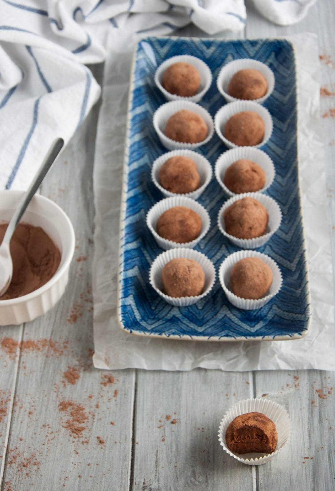 A rectangular blue plate holds ten avocado chocolate truffles, each nestled in a white paper cup. A bowl of cocoa powder and a spoon sit nearby. On the wooden surface, one truffle reveals a tempting bite.