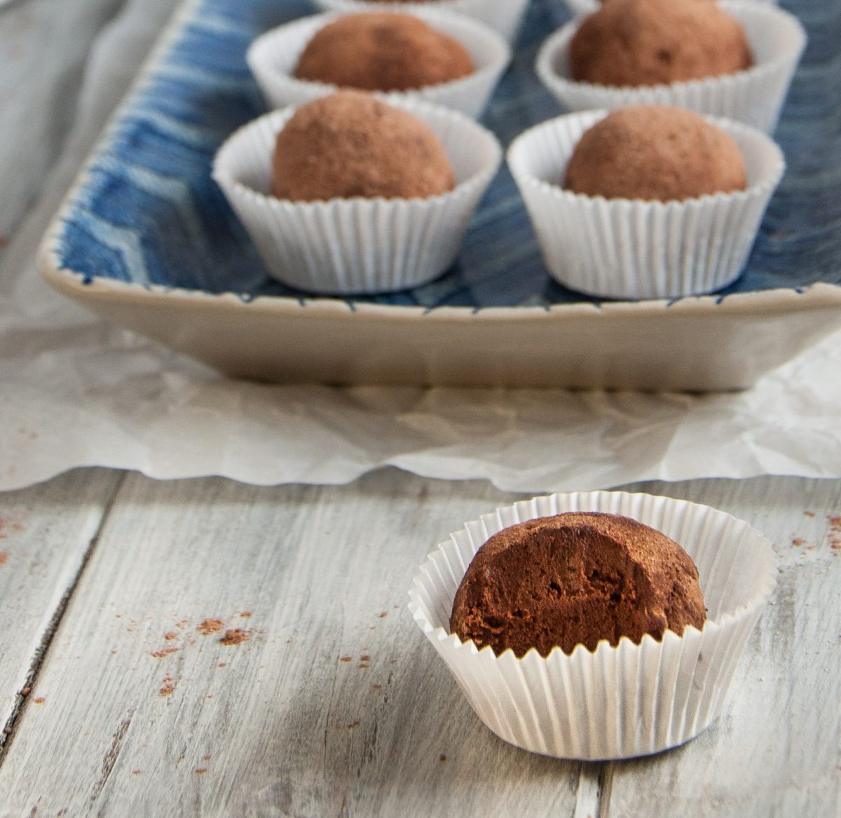 Rich avocado chocolate truffle nestled in a paper cup with a bite taken out of it on a rustic wooden surface, with a tray showcasing more truffles in the background.