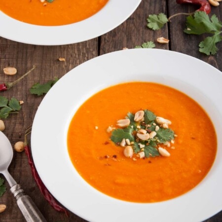 Two bowls of orange soup garnished with peanuts and cilantro on a wooden table, surrounded by cilantro leaves and red peppers.