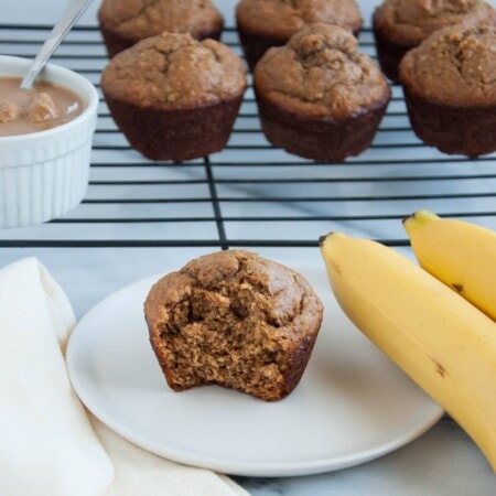 Banana muffin with a bite taken out on a white plate, surrounded by ripe bananas and more muffins on a cooling rack.