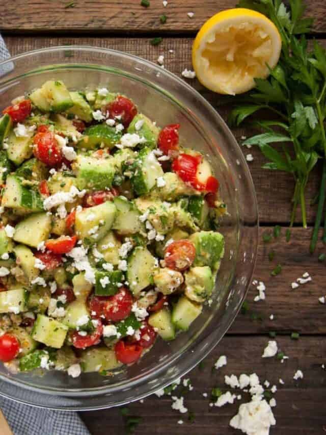 tomato avocado cucumber salad on a wood background with lemon, feta and parsley next to it