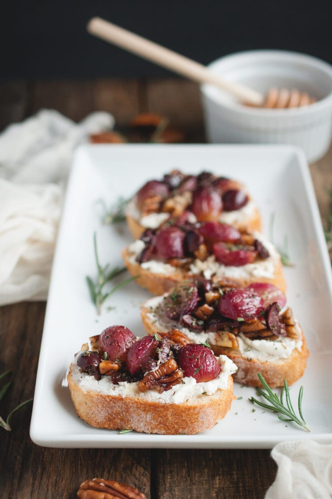 Plate with crostini topped with roasted grapes, goat cheese, rosemary, and kalamata olives with honey in background