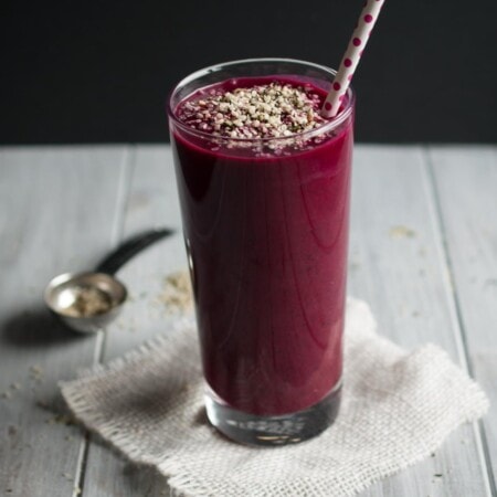 A tall glass of purple smoothie topped with seeds, placed on a white napkin. A metal spoon with seeds is in the background. A polka dot straw is in the glass.