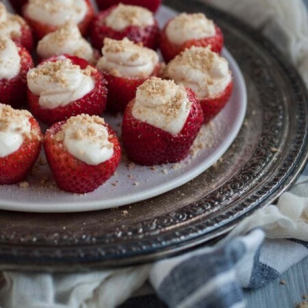 Strawberries filled with cream cheese and topped with crumbs are arranged on a white plate with a decorative silver tray underneath.