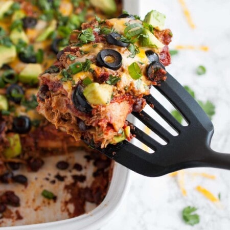 A slice of Mexican casserole with cheese, olives, green onions, and avocado is lifted from a baking dish with a black spatula. The surface is garnished with cilantro.