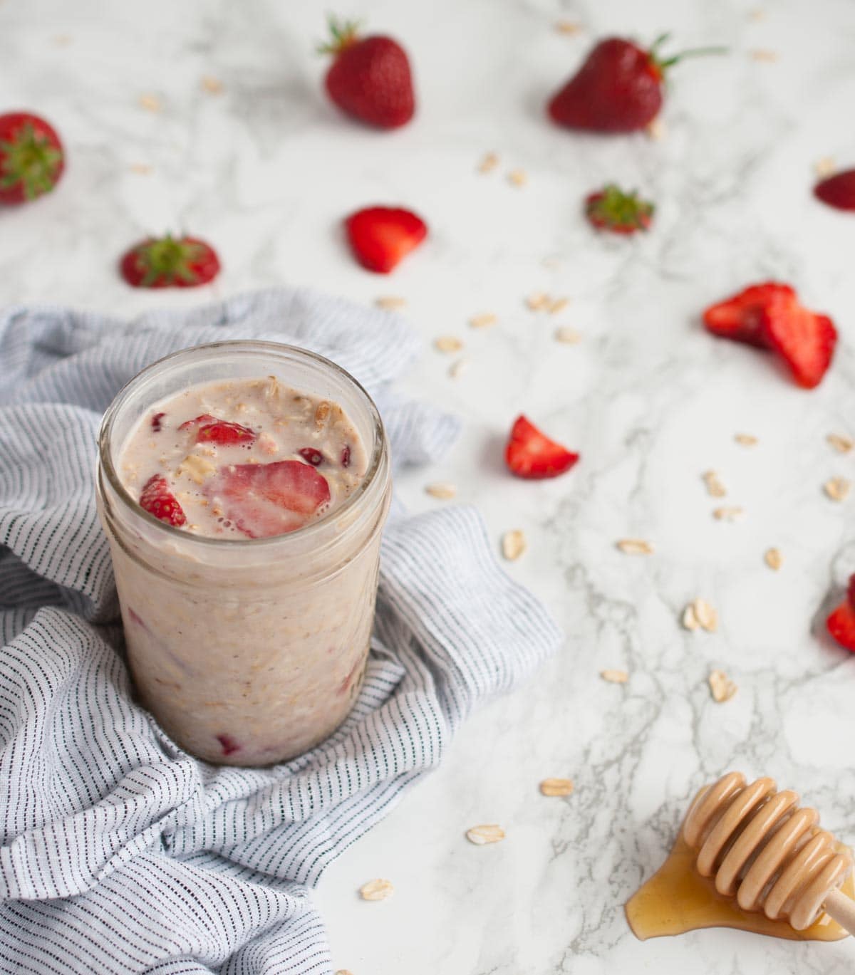 side angle of a jar of overnight oats on a marble backdrop with oats, sliced strawberries sprinkled on it next to a honey dipper 