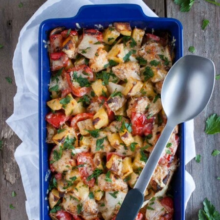 A blue baking dish contains a baked vegetable and cheese casserole, garnished with parsley. A serving spoon rests on top.