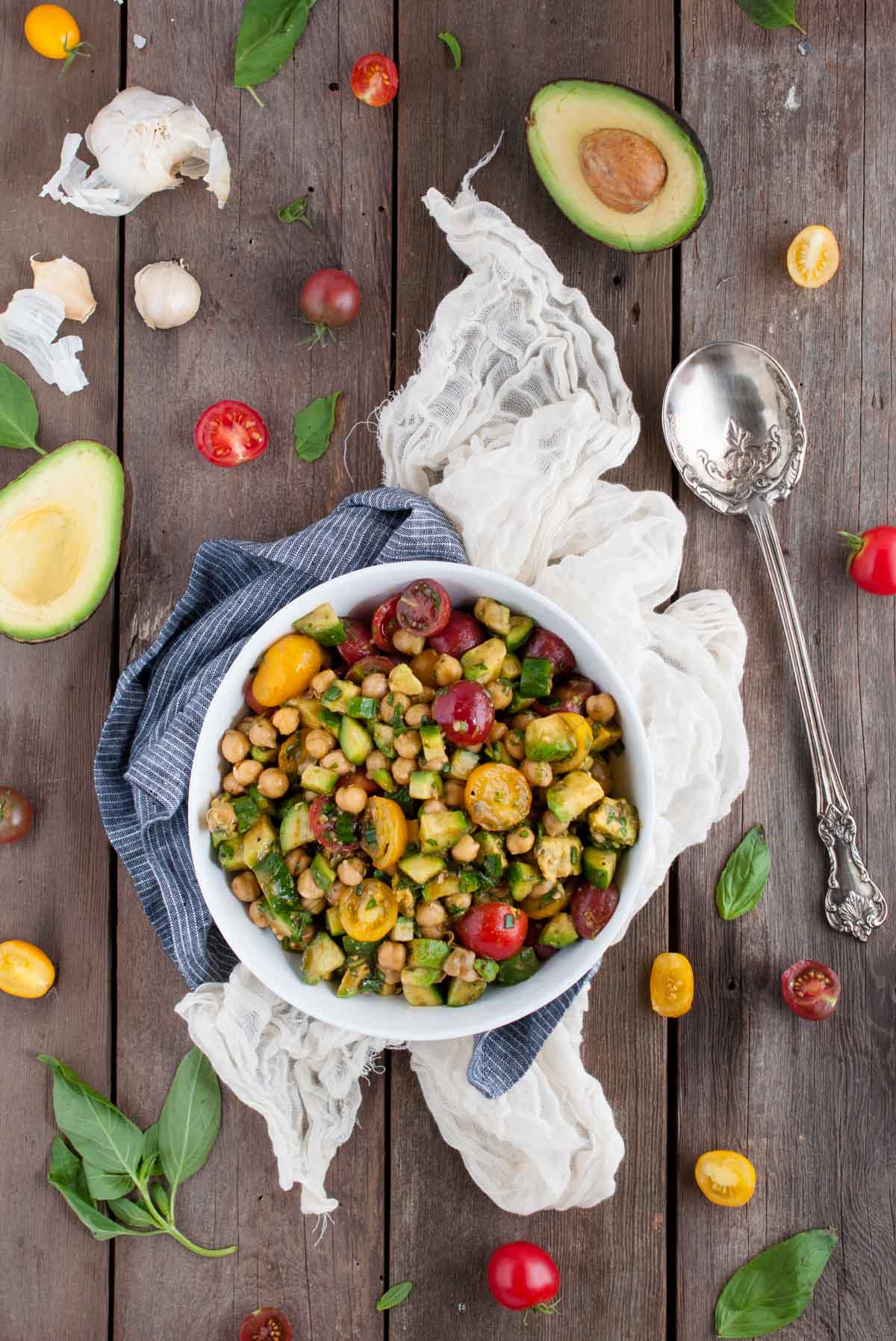 chickpea salad from overhead angle in a white bowl on a wood background with salad ingredients sprinkled around it and a serving spoon