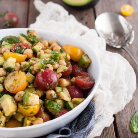 A bowl of salad with grape tomatoes, chickpeas, cucumber, avocados, and herbs on a wooden table with a spoon and scattered cherry tomatoes.