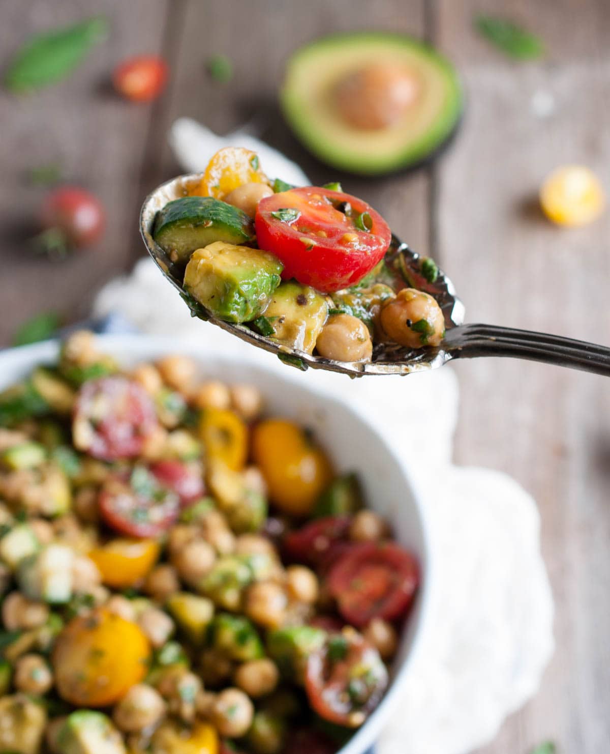 serving spoon with tomato avocado cucumber chickpea salad being held up with full bowl of more salad in background