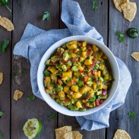 A bowl of mango salsa with diced mango, red pepper, avocado, cilantro, and red onion on a wooden table, surrounded by tortilla chips and lime halves.
