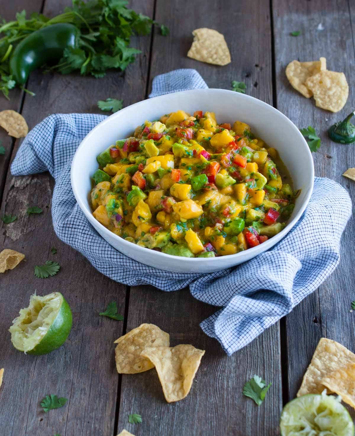side angle of bowl of fruity salsa with a squeezed lime, chips and other ingredients on the side on a wood background