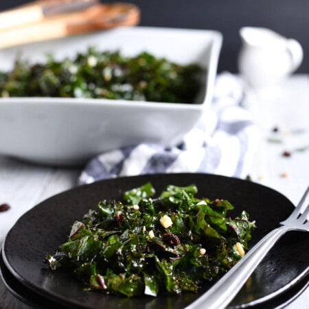 A plate of kale salad with nuts and raisins, accompanied by a fork. In the background, a larger bowl of the same salad and a small pitcher are visible.