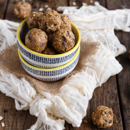 A bowl of round, homemade energy balls with oats and chocolate chips on a wooden table, surrounded by a white cloth and scattered oats.