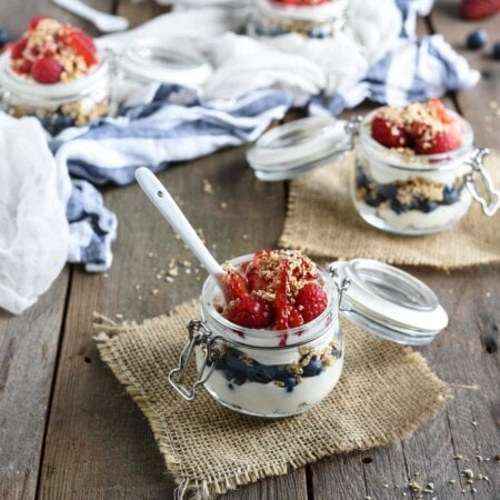 Small glass jars with yogurt, granola, strawberries, and raspberries on wooden table. Blueberries scattered nearby. Cloth and additional jars in the background.