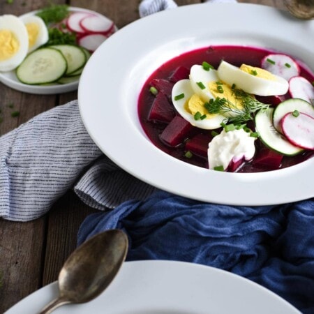 A bowl of chilled beet soup garnished with sliced boiled eggs, radishes, cucumbers, and dill, served on a wooden table with a spoon and a striped cloth napkin.