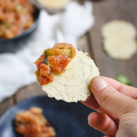 A hand holding a round cracker topped with chunky salsa, with more salsa and crackers in the background.