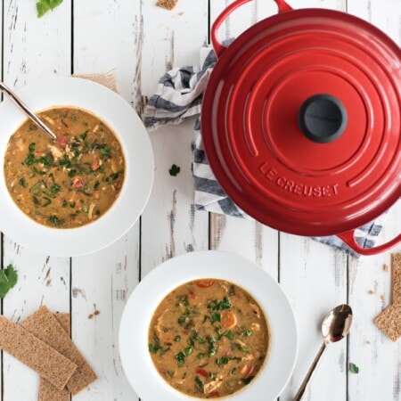Two bowls of vegetable soup with spoons on a white wooden table, alongside a red pot and cracker pieces. Parsley leaves are scattered around.