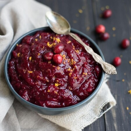 Bowl of vibrant red cranberry sauce garnished with orange zest and whole cranberries, with a silver spoon resting on a beige cloth.