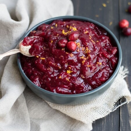 A bowl of cranberry sauce with whole cranberries and a spoon, placed on a textured cloth.