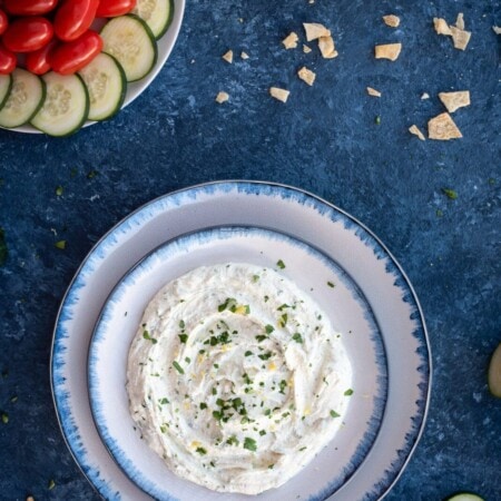 A bowl of creamy dip garnished with herbs, surrounded by sliced cucumbers, cherry tomatoes, and a bowl of pita chips on a blue surface.