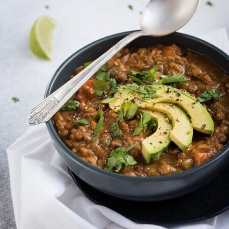 Bowl of lentil stew topped with sliced avocado and herbs, placed on a white cloth. A spoon rests on the bowl, with a lime wedge nearby.
