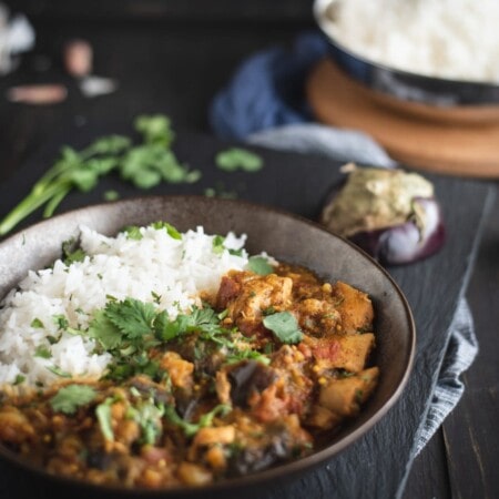 A bowl of white rice and vegetable curry garnished with cilantro, placed on a dark slate surface. An eggplant and a dish of rice are visible in the background.