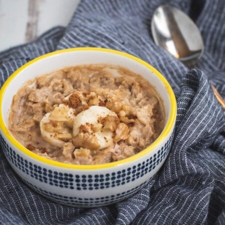 Bowl of oatmeal topped with banana slices and walnuts, set on a striped cloth with a spoon beside it.