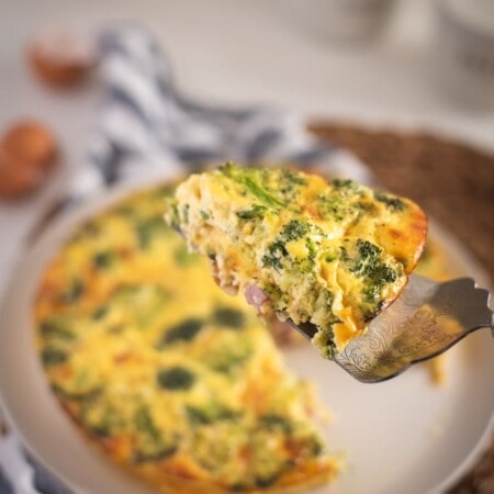 A slice of broccoli and cheese quiche is being lifted from a whole quiche on a plate.