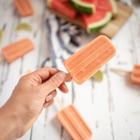 A hand holds a peach-colored popsicle. More popsicles and watermelon slices on a wooden board are in the background.