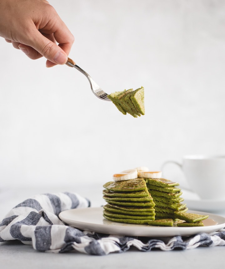 hand holding bite of green pancakes over plate of pancakes