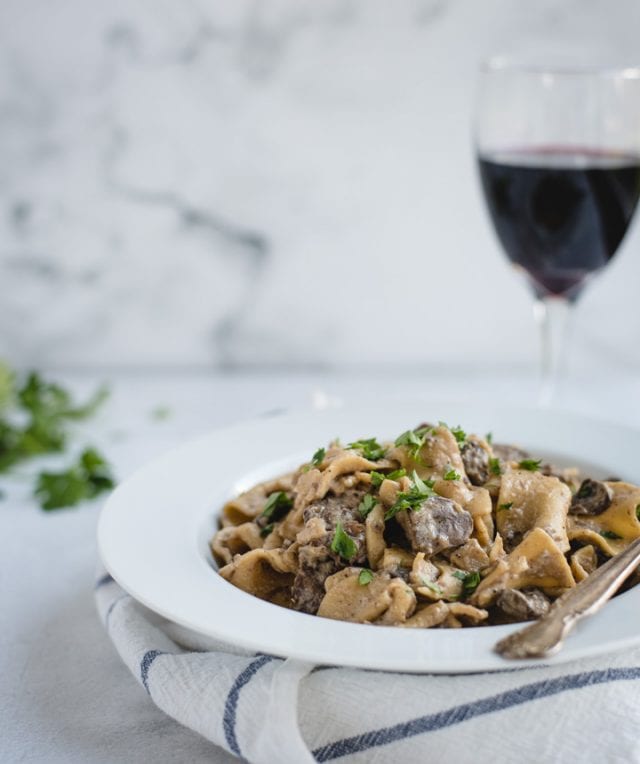 Bowl of beef stroganoff with glass of red wine on white background