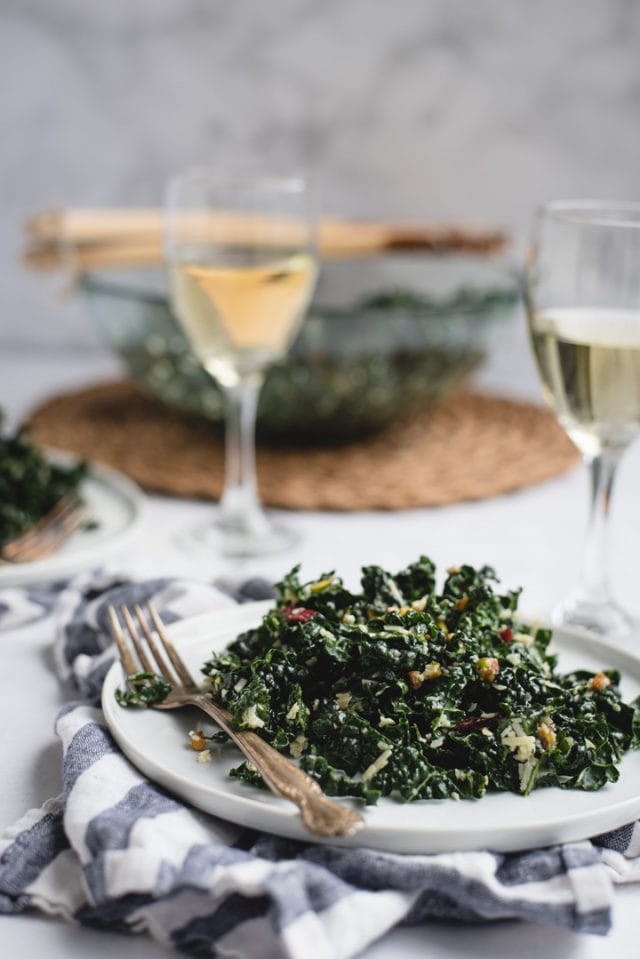 side view picture of kale salad on a plate with wine and big salad bowl in background