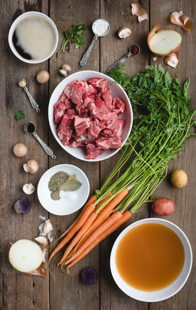 irish beef stew ingredients spread out on a wood background (cubed beef, carrots, broth, spices, etc.)