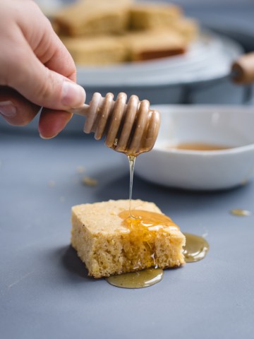 hand holding honey dipper drizzling honey onto a piece of cornbread