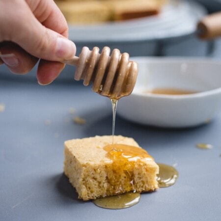 A hand drizzles honey over a square piece of cornbread on a gray surface. A bowl of honey and more cornbread pieces are in the background.