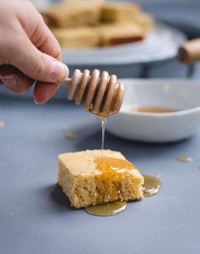 Hand holding a honey dipper drizzling honey onto a piece of cornbread