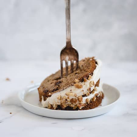 A slice of frosted cake with nuts, placed on a white plate. A fork is inserted into the center.